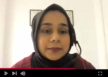 Individual wearing a hijab participating in a video call, with a plain white background and a framed picture hanging on the wall. There is a play button overlay at the bottom center, indicating that this is a still from a video.